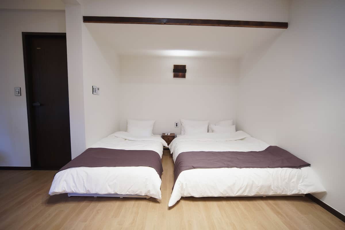 A room with two beds, a wooden floor, a door, and a small wall-mounted light fixture.