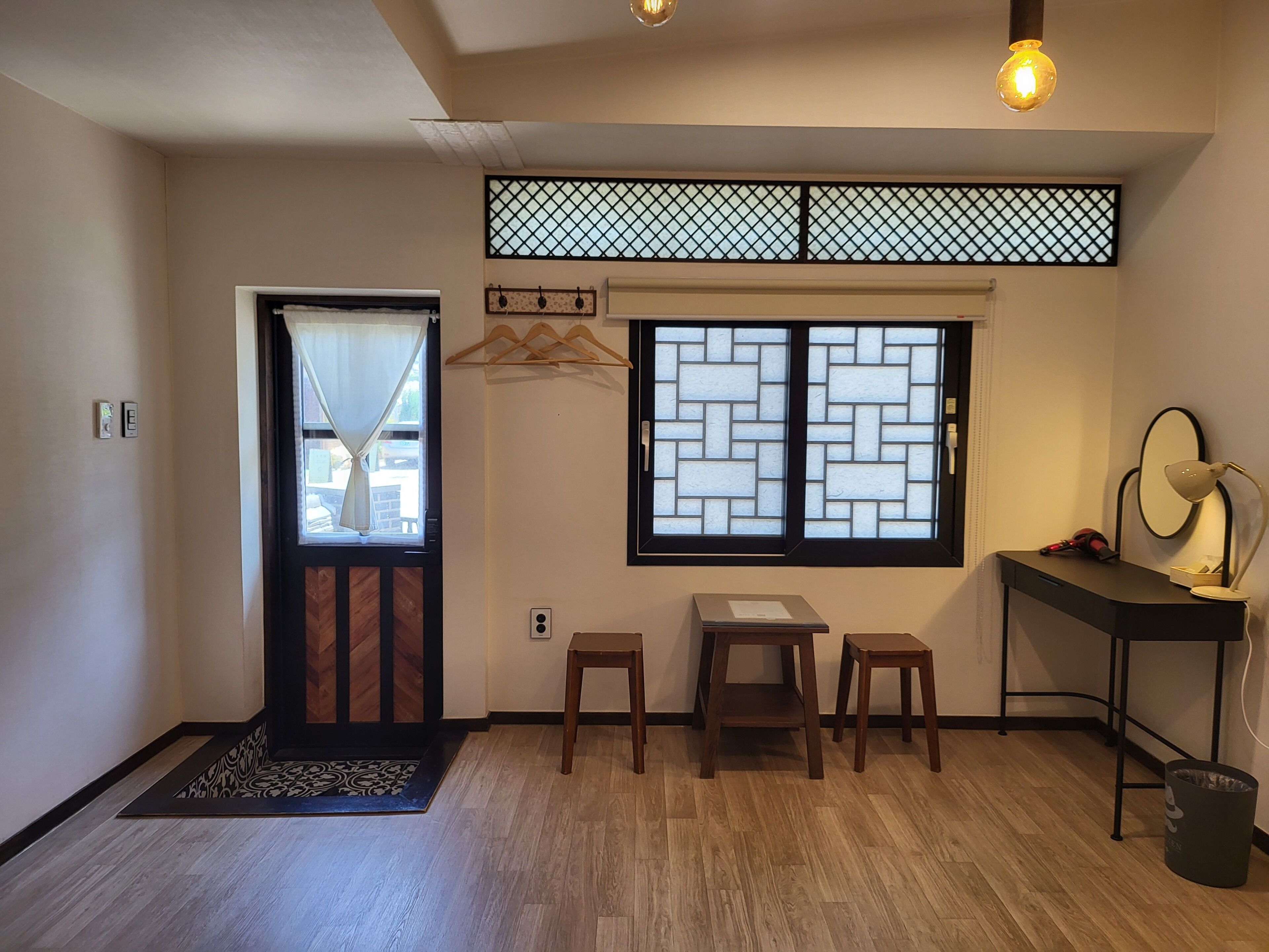 A room with a wooden floor, a desk with a lamp, a chair, a small table, a hanging light fixture, and a window with a decorative screen.