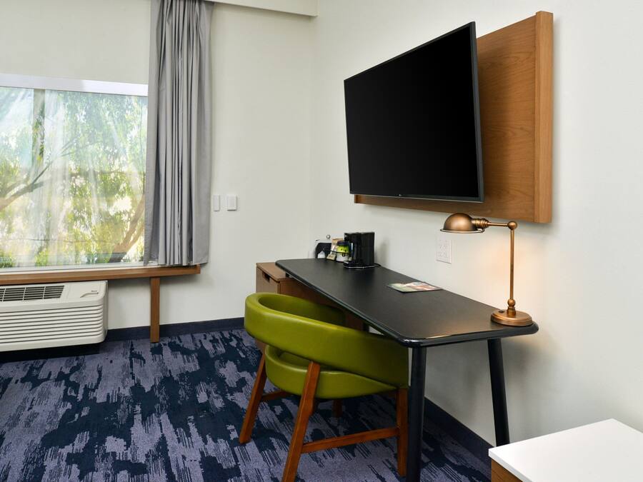 A hotel room with a desk, a green chair, a flat-screen TV mounted on the wall, a window with a view of trees, and a ceiling fan.