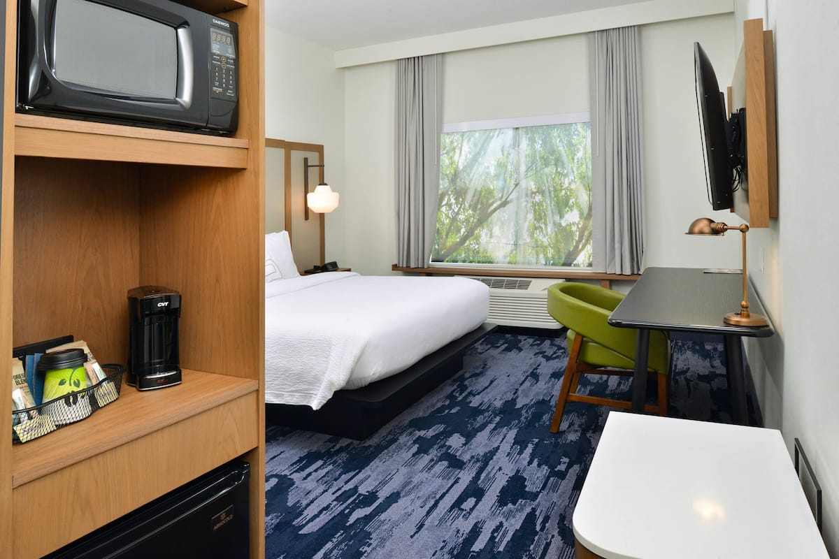 A hotel room with a bed, a desk, a chair, a TV, a microwave, a coffee maker, and a window with a view of trees.