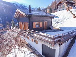 Una cabaña con techo inclinado, contraventanas de madera y una antena parabólica. Está rodeada de nieve y montañas al fondo.