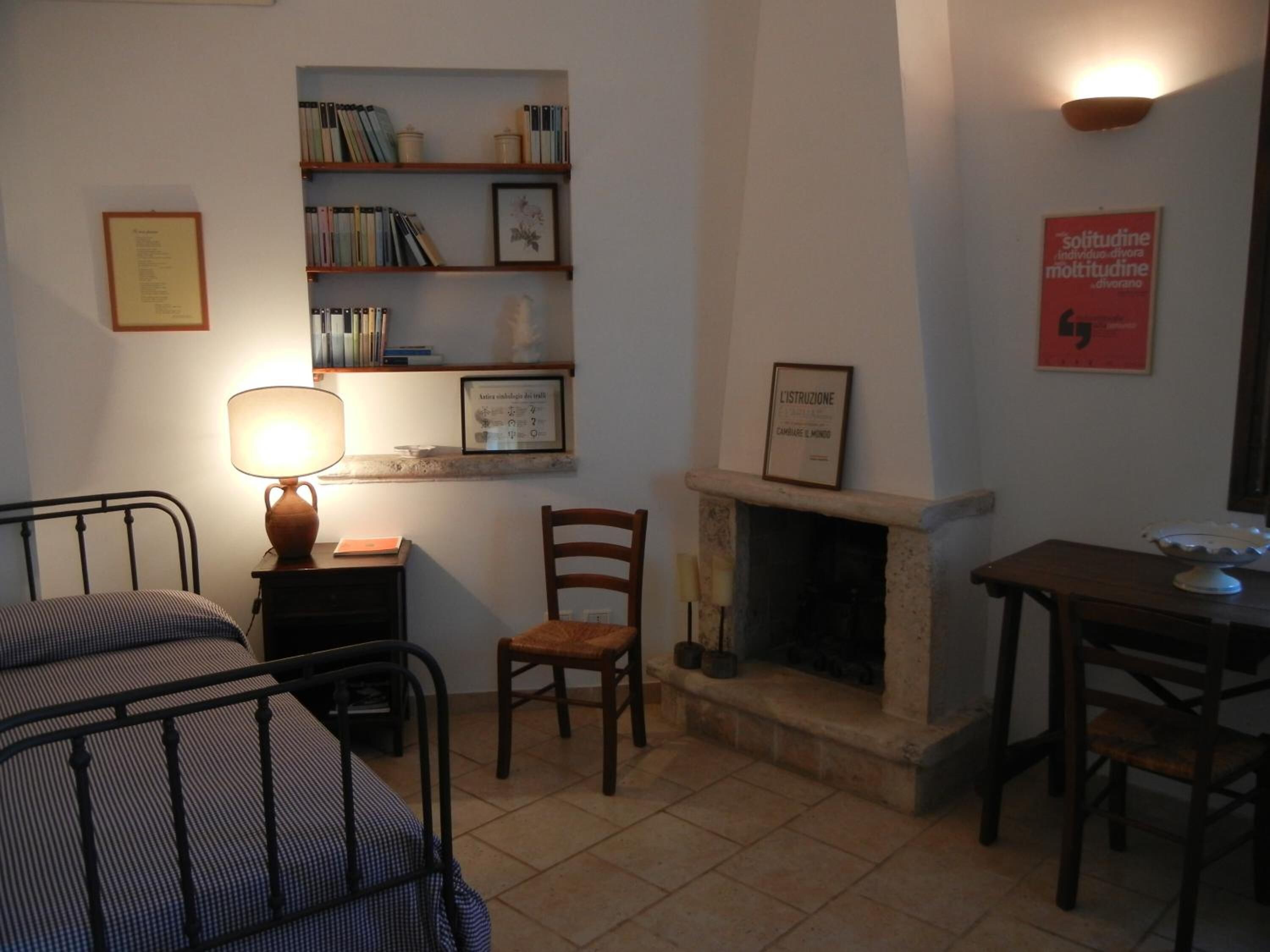 A cozy room with a bed, a chair, a small table, a bookshelf, a fireplace, and a wall-mounted lamp.