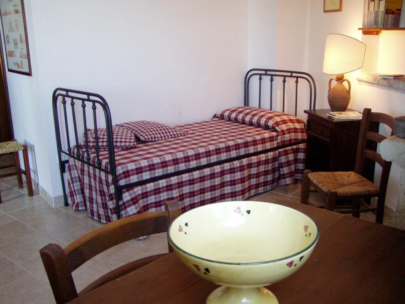 A small, cozy room with two metal beds, a wooden chair, a table with a bowl, and a lamp on a nightstand.