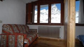 A room with a wooden floor, a patterned sofa, a window with a view of snow-capped mountains, and a radiator. - Room Apartment Luxury Duplex view Matterhorn skis - SAUNA - WIFI (Breuil-Cervinia)