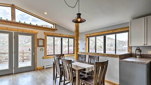 Un comedor amplio con una mesa y sillas de madera, ventanas grandes que ofrecen vistas al exterior, y una zona de cocina con armarios y una encimera.