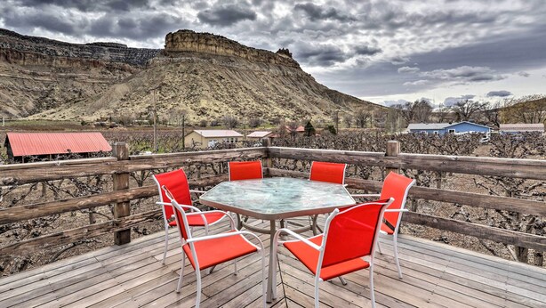 Una terraza de madera con una mesa de vidrio y sillas rojas, con vistas a un viñedo y una montaña.