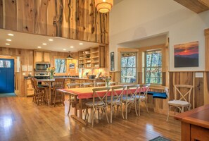 Une cuisine spacieuse avec des meubles en bois, un coin repas agrémenté d’une nappe rouge et de grandes fenêtres offrant une vue sur les arbres.
