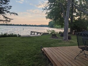 A wooden deck with a bench and a table, overlooking a lake with a dock and a fire pit.