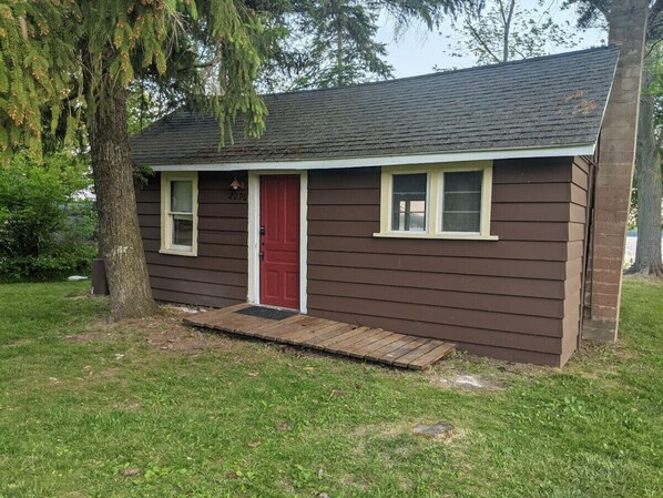 A small, single-story building with a red door and two windows, surrounded by grass and trees.