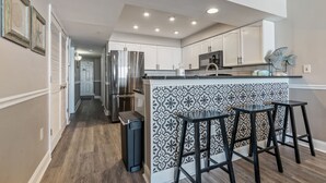 Een moderne keuken met witte kastjes, een roestvrijstalen koelkast en een decoratieve spatwand. Er zijn twee zwarte barstoelen en een afvalbak.