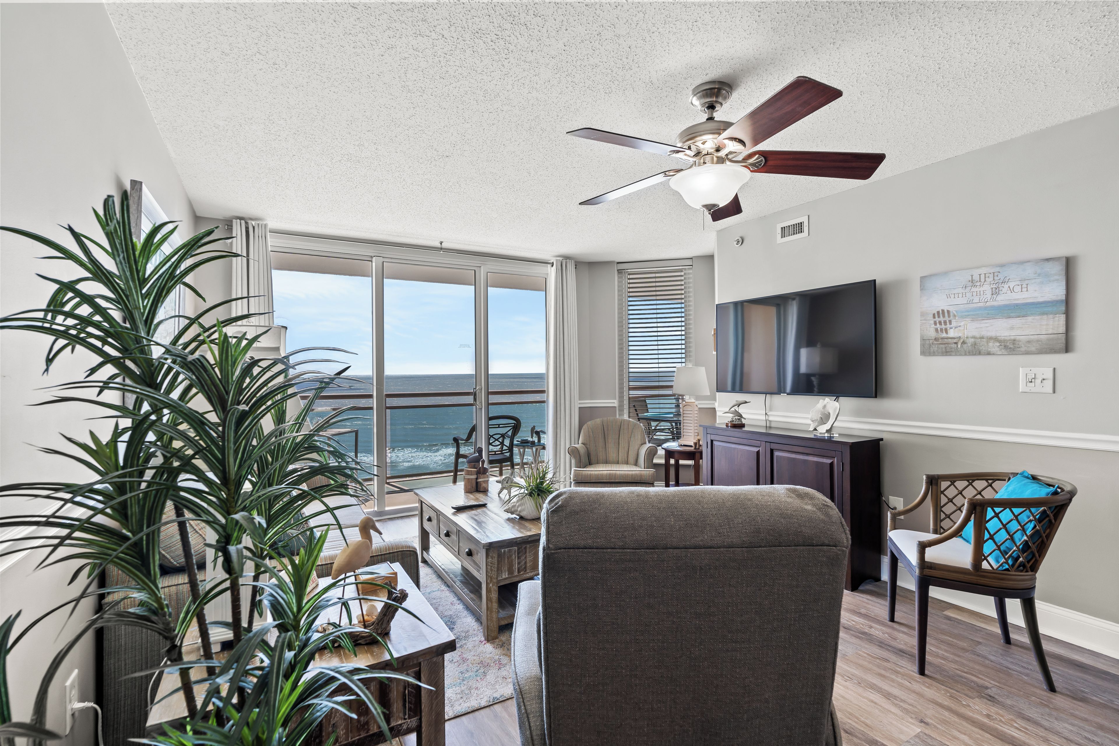 A spacious living room with a large window overlooking the ocean, a ceiling fan, and a flat-screen TV mounted on the wall.