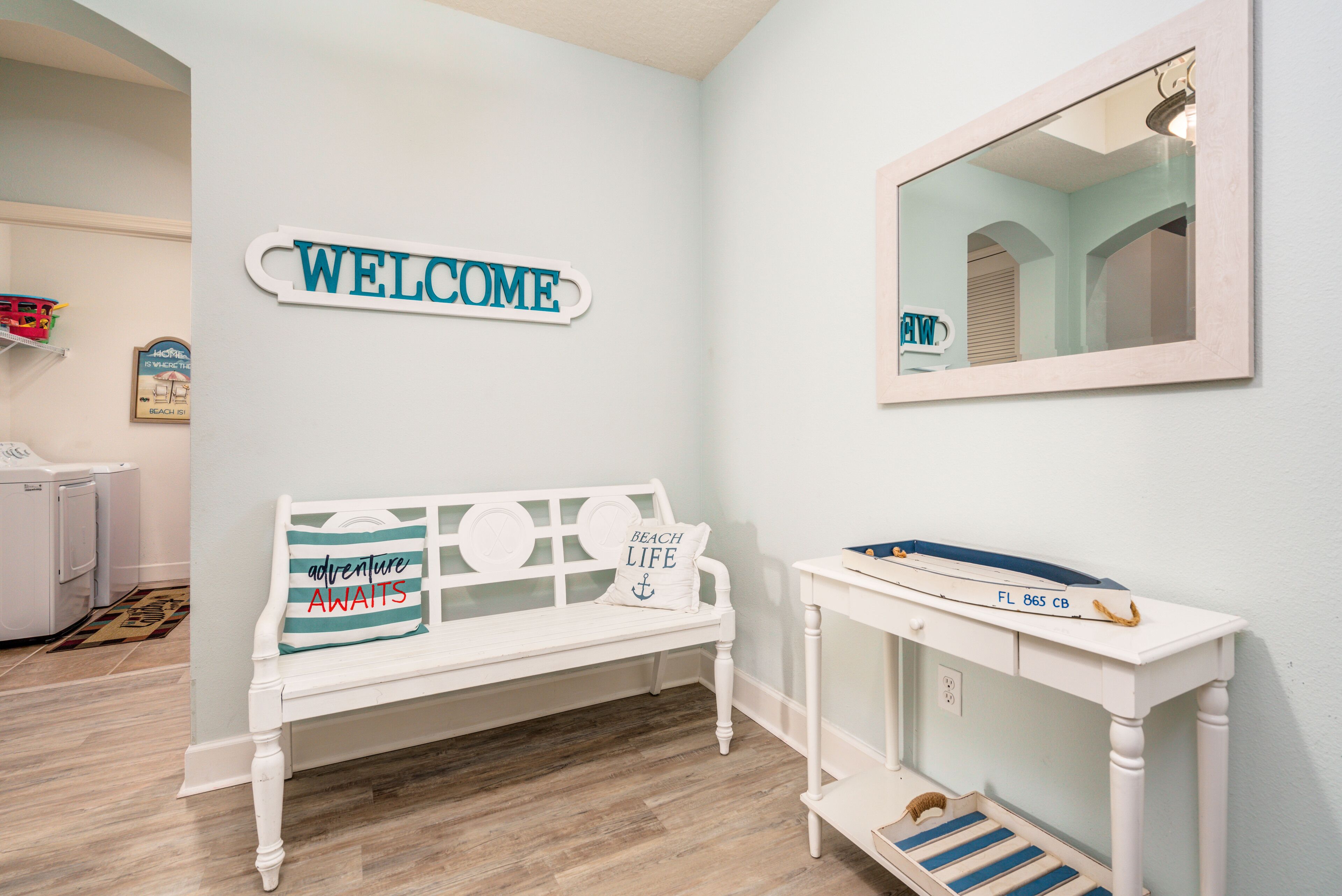 A room with a white bench, a welcome sign, a mirror, and a small table with a tray.