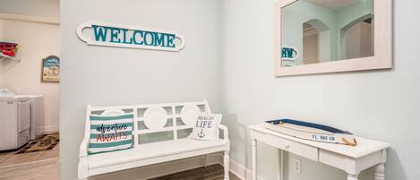 A room with a white bench, a welcome sign, a mirror, and a small table with a tray.