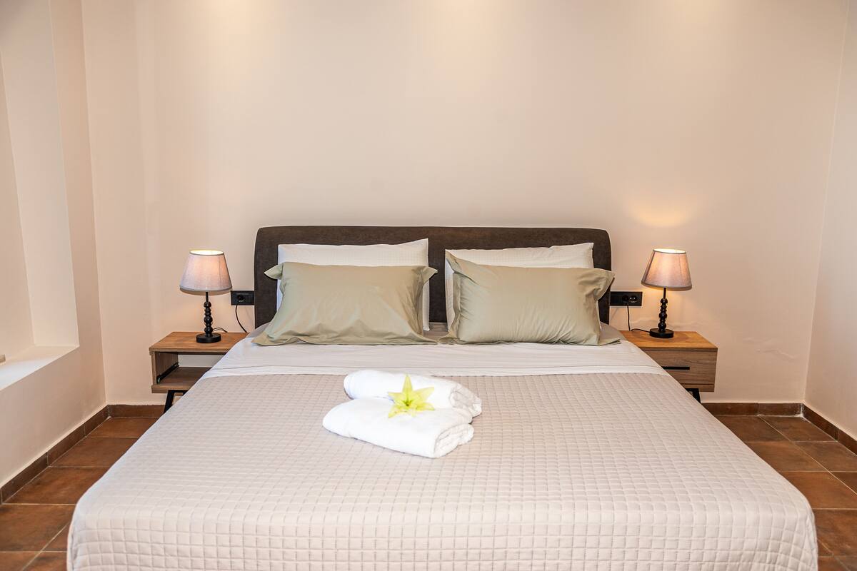A neatly made bed with a white quilt, two pillows, and a towel with a flower. There are two bedside lamps on wooden nightstands.