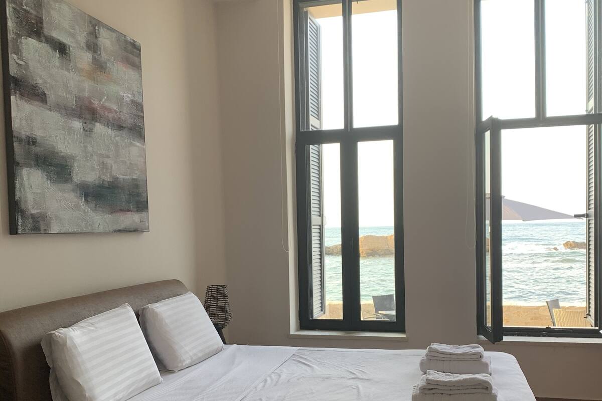 A bedroom with a large bed, two windows with ocean views, and a painting on the wall.