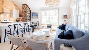 Eine moderne Küche mit Backsteinwand, weißen Schränken und einer Kochinsel. Ein Essbereich mit rundem Tisch und Stühlen sowie ein Wohnzimmer mit blauem Sofa und großem Fenster.