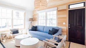 Ein Zimmer mit einem blauen Sofa, einem runden Couchtisch, Korbstühlen, einer Lampe und einer dunklen Holztür.