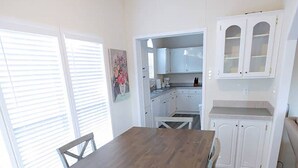 Un coin repas avec une table en bois et des chaises blanches, une cuisine avec des meubles blancs et une fenêtre équipée de stores.