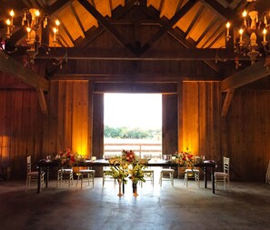 Tunnelmallinen, maalaishenkinen ruokailutila, joka on sisustettu vanhan navetan tyyliin. Siinä on puiset kattopalkit, koristeelliset kattokruunut ja suuri ruokapöytä, joka on katettu valmiiksi.