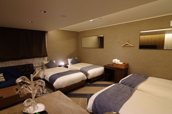 Habitación de hotel con dos camas, una mesita de noche con lámpara, un espejo, perchero y una pequeña mesa con un cesto de artículos de aseo.
