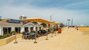 A seaside restaurant with outdoor seating, a white building, and a sandy beach in the background.
