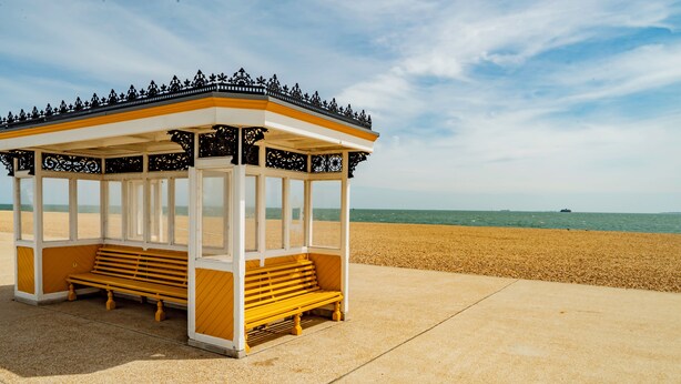 A beach gazebo with a bench, a clear view of the ocean, and a sandy beach.