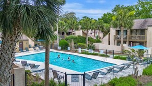 Una piscina rodeada de palmeras y sillas de descanso, con una valla y un cartel que indica que no se permiten recipientes de vidrio en el área de la piscina.