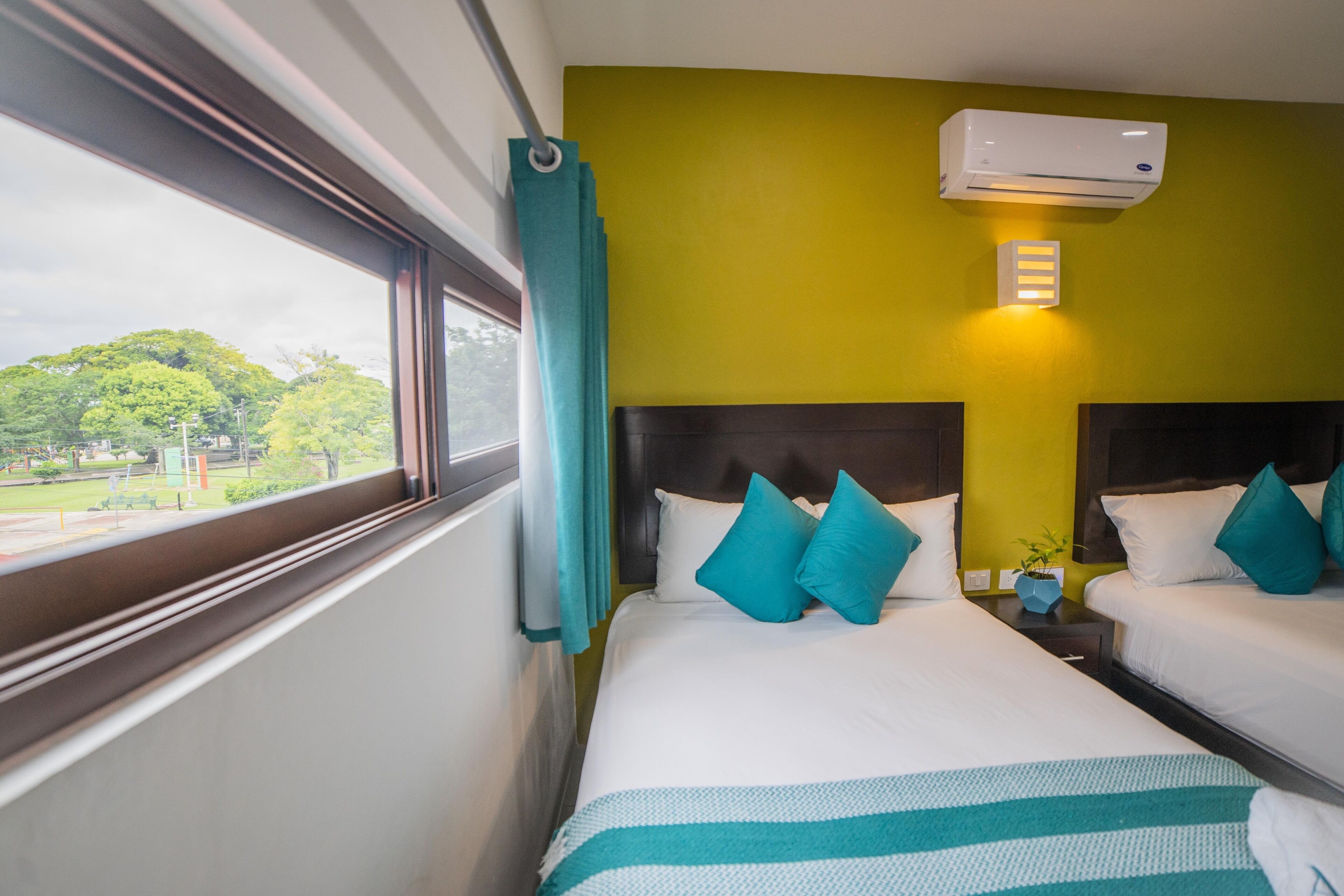 A hotel room with two beds, a window with curtains, a wall-mounted air conditioner, and a small plant on the nightstand.