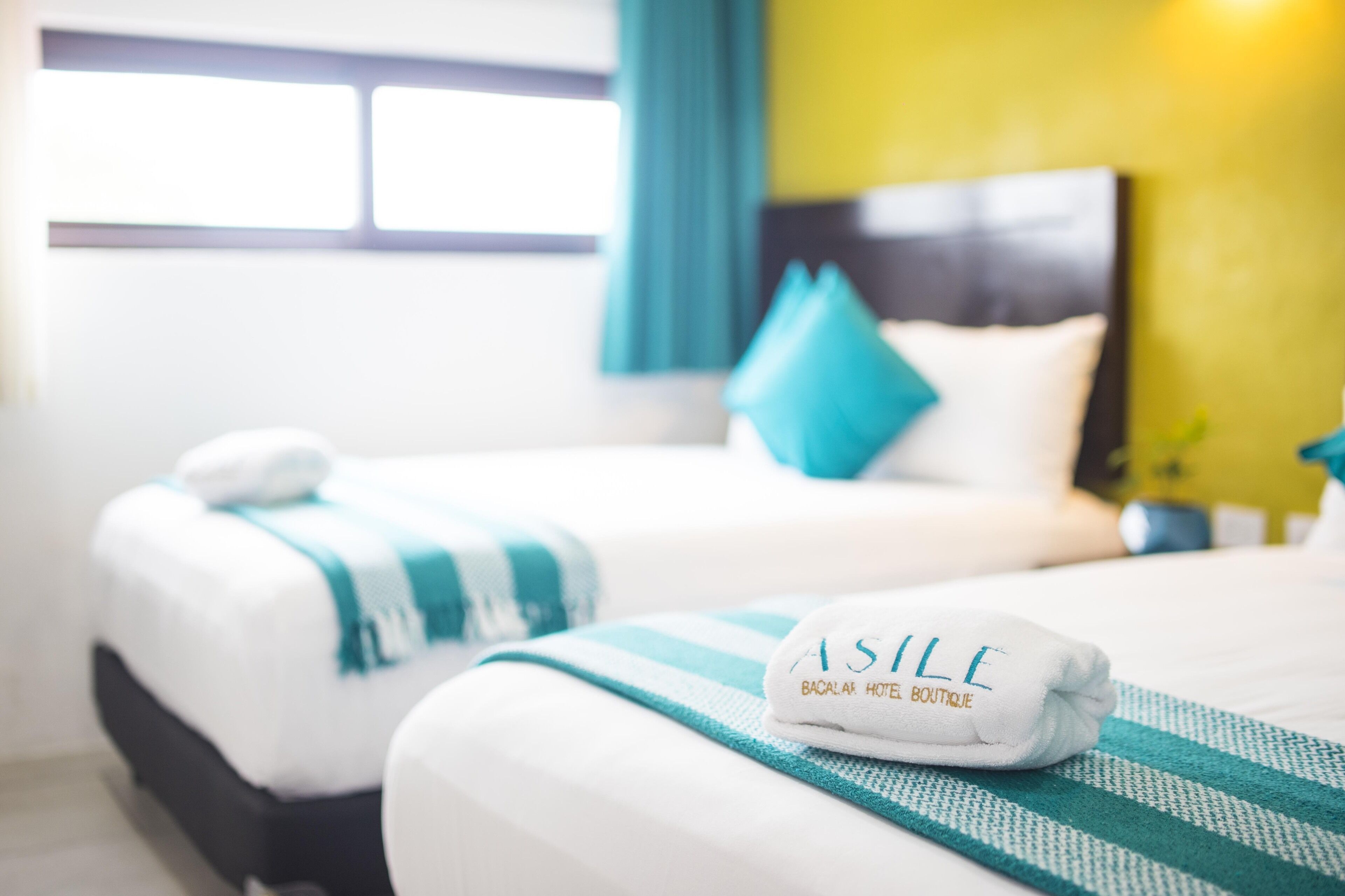 A hotel room with two beds, each with a white and teal striped blanket, and a white towel with "ASILE" on it.