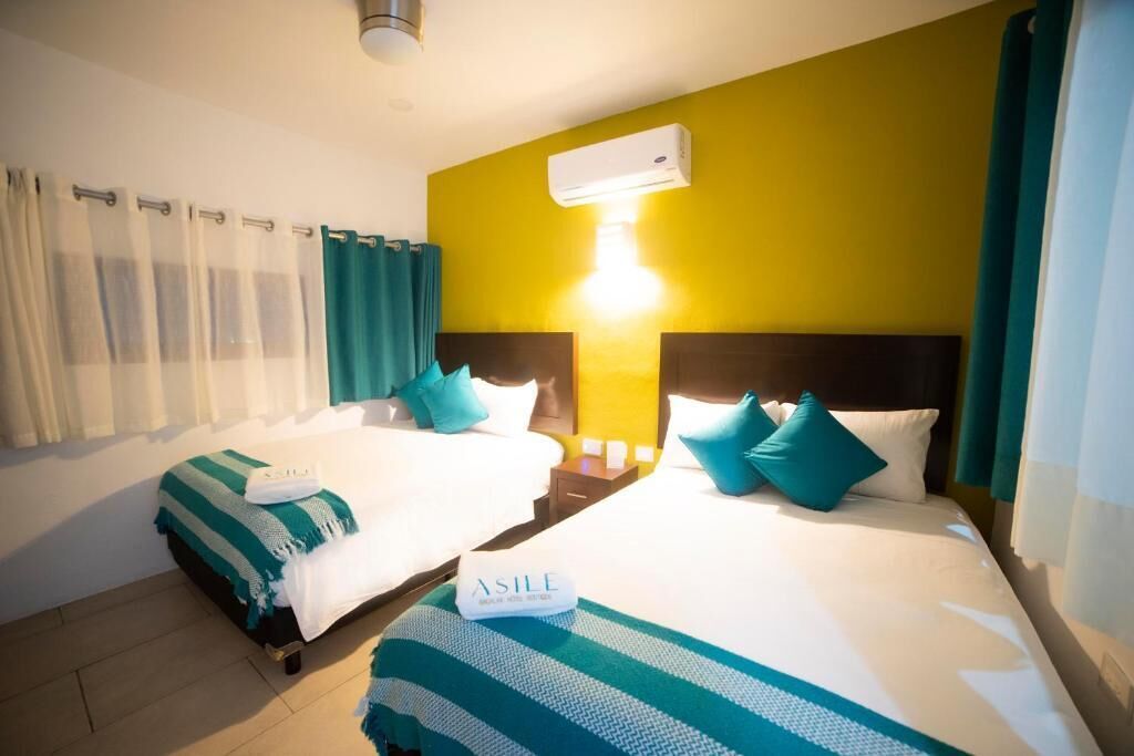 Habitación de hotel con dos camas, una pared de acento amarilla, un equipo de aire acondicionado y una mesita de noche con lámpara.