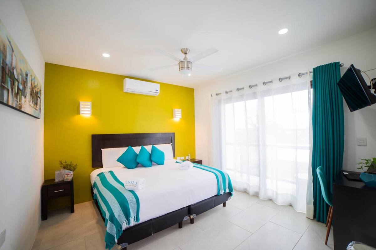 Habitación de hotel con cama, escritorio, silla, ventilador de techo y ventana con cortinas.