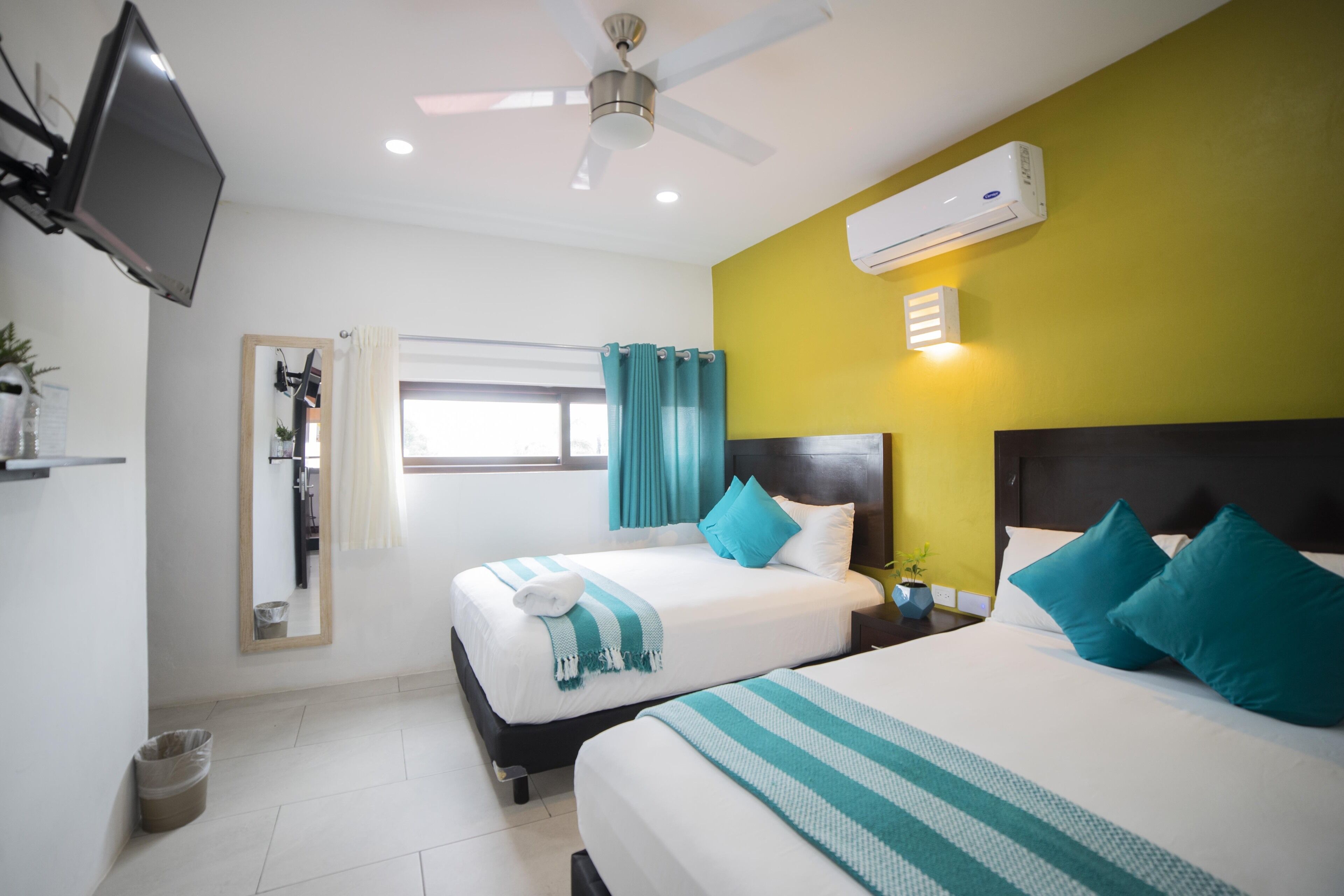 A hotel room with two beds, a ceiling fan, an air conditioning unit, a mirror, a window with curtains, and a small plant on the nightstand.
