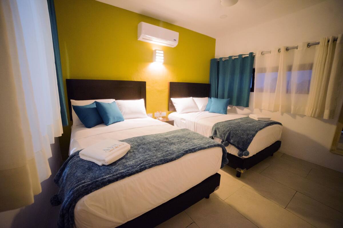 Una habitación de hotel con dos camas, cada una con cabecero, ropa de cama blanca y almohadas azules. Se observa un aire acondicionado instalado en la pared y una ventana con cortinas.