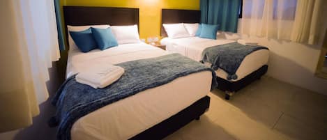 A hotel room with two beds, each with a headboard, white bedding, and blue pillows. There is a wall-mounted air conditioner and a window with curtains.