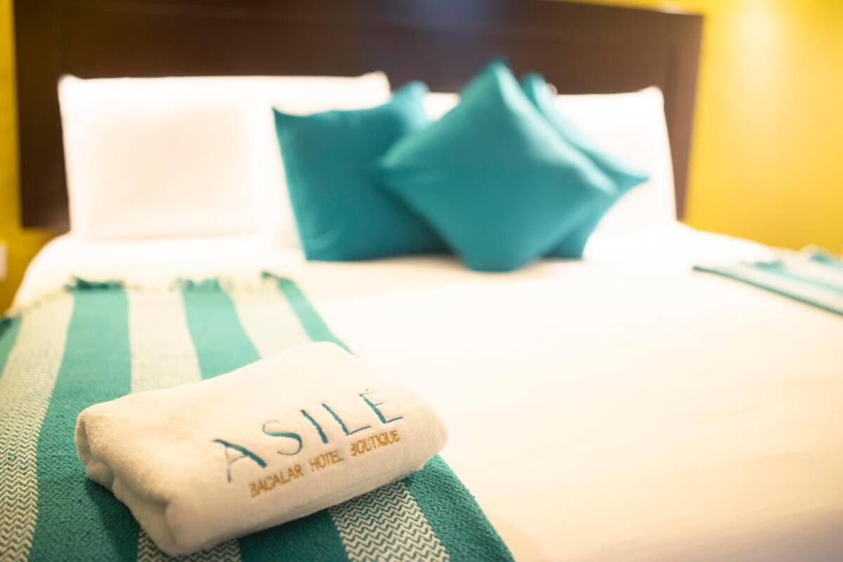 Una cama bien tendida con una toalla doblada que dice 'ASILE' y una manta verde a rayas.