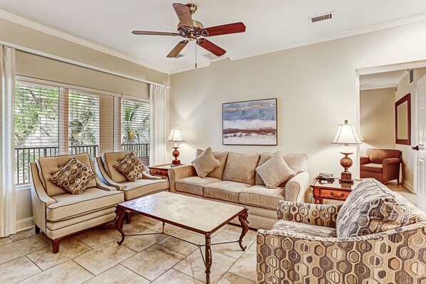 A living room with a beige sofa set, patterned armchair, and a coffee table. There is a ceiling fan, a painting on the wall, and a window with blinds.