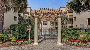 Ein begrenzter Eingangsbereich mit einer Pergola, Palmen und einem Ziegelweg, der zu einem Gebäude mit mehreren Balkonen führt.