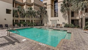 A swimming pool with a poolside area, surrounded by a multi-story building with balconies and palm trees.