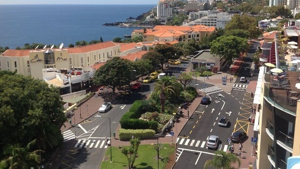 A coastal cityscape with a prominent road intersection, buildings with red-tiled roofs, and a view of the sea. - Cosy  Apartment at Lido with Pool (Funchal)