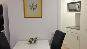 A small dining area with a white table and two black chairs, a framed botanical print on the wall, and a kitchen with a microwave and cabinets. - Cosy  Apartment at Lido with Pool (Funchal)