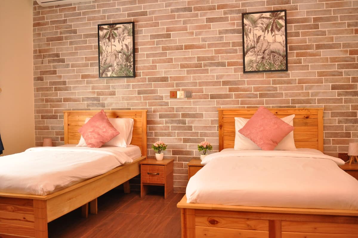 A room with two wooden beds, each with a wooden headboard, white bedding, and pink pillows. There are small wooden nightstands with lamps and potted plants. The wall is brick with two framed pictures.