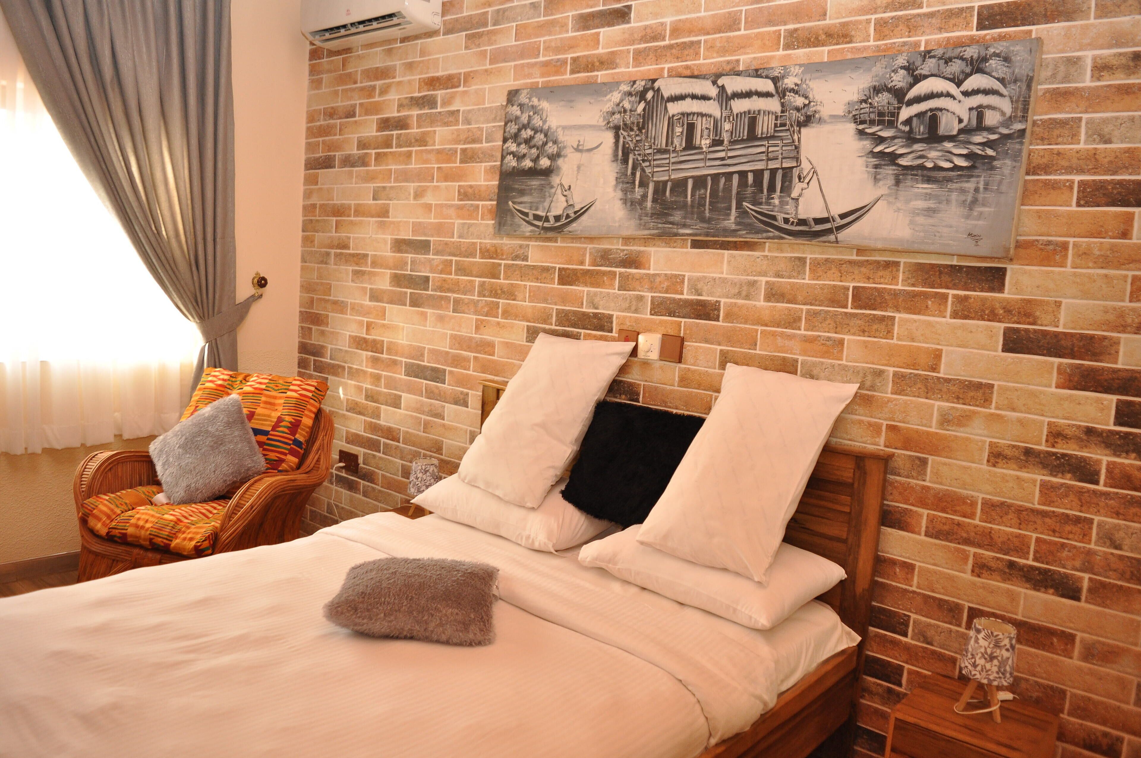 A bedroom with a brick wall, a bed with pillows, a chair with a cushion, and a painting of a boat on a river.
