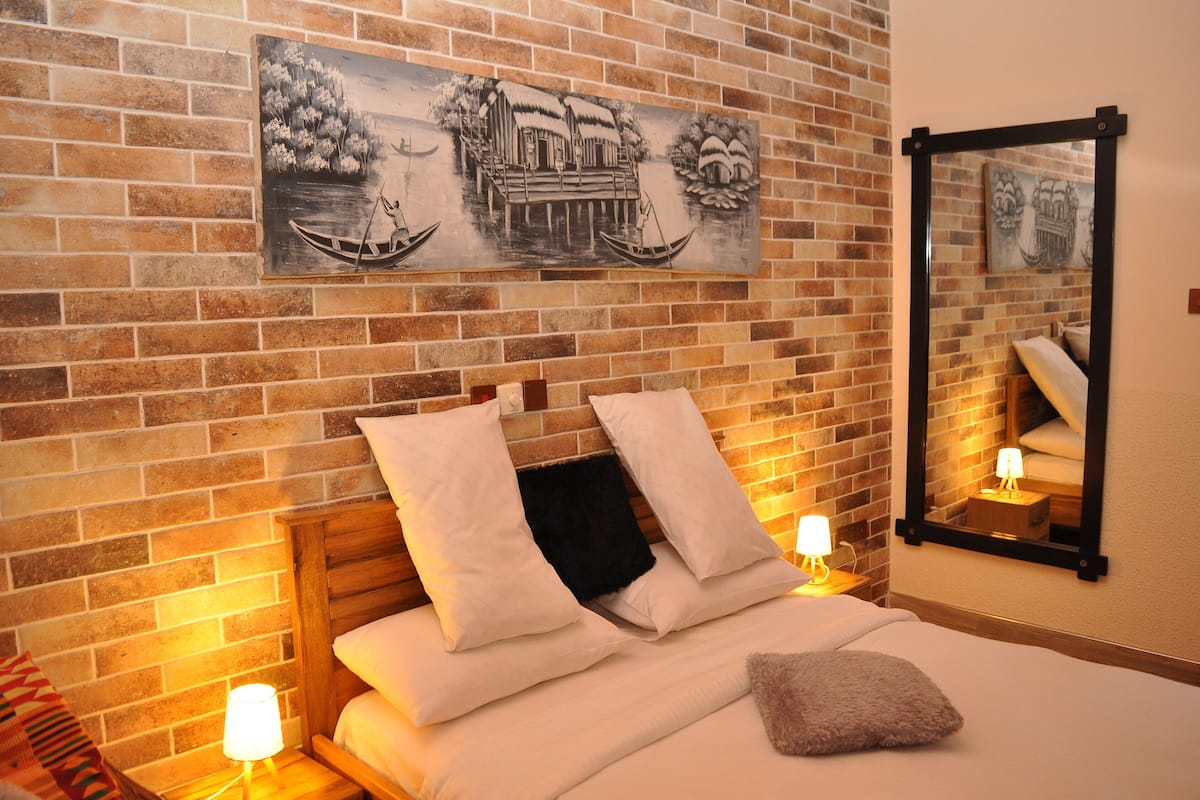 A hotel room with a brick wall, a bed with white bedding, two pillows, a nightstand with a lamp, and a painting of a traditional scene.