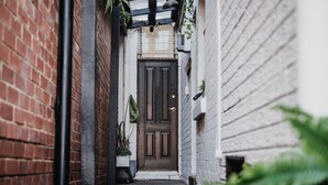 Une ruelle étroite, avec une porte en bois sombre au fond, bordée de murs en briques et de plantes en pot.