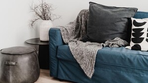 Un salon moderne avec un canapé de couleur turquoise, un plaid gris et un coussin noir et blanc. On y trouve un pouf rond en cuir et un vase blanc orné de branches, posés sur une table d’appoint.
