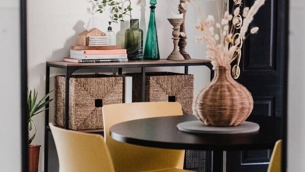 Un coin repas avec une table ronde, deux chaises jaunes, une bibliothèque remplie de livres et d’objets décoratifs, et une porte en arrière-plan.