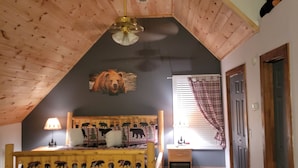 Un dormitorio con techo inclinado, paredes de madera, una cama con ropa de cama temática de animales, un ventilador de techo y una ventana con cortinas.