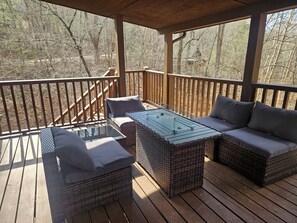 Eine überdachte Terrasse im Freien mit Rattanmöbeln, darunter ein Sofa und zwei Sessel, sowie ein Tisch mit Glasplatte.