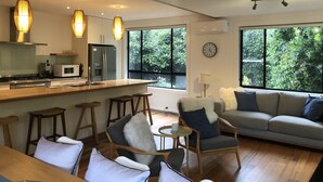A modern kitchen with a central island, bar stools, and a dining table. There is a living area with a sofa and chairs, and large windows offering a view of greenery. - Heart of Red Hill (Red Hill)