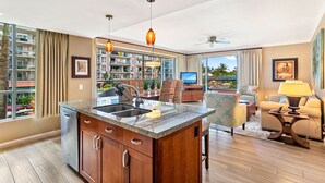 A modern kitchen with a granite countertop, wooden cabinets, and a built-in dishwasher. There is a dining area with a table and chairs, and a living room with a sofa and a TV. The room has large windows with a view of the outside. - Maui Resort Rentals: Honua Kai Konea 239 - Upgraded Ocean View 2BR, Deluxe Corner Suite (Lahaina)